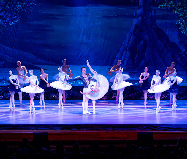 A stage scene during a performance of the ballet Swan Lake, with 13 female dancers in white or black tutus and one male dancer.