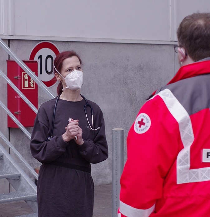 A female doctor wearing a mask covering her mouth and nose and protective clothing talks to a German Red Cross worker